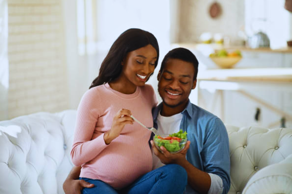 Joyful Black couple enjoying a fresh vegetable salad at home, embracing healthy nutrition to support their IVF pregnancy and promote maternal well-being